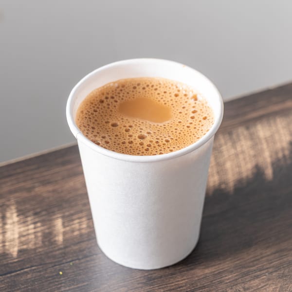 A white paper cup filled with frothy, light brown chai tea sits on a dark wooden surface.