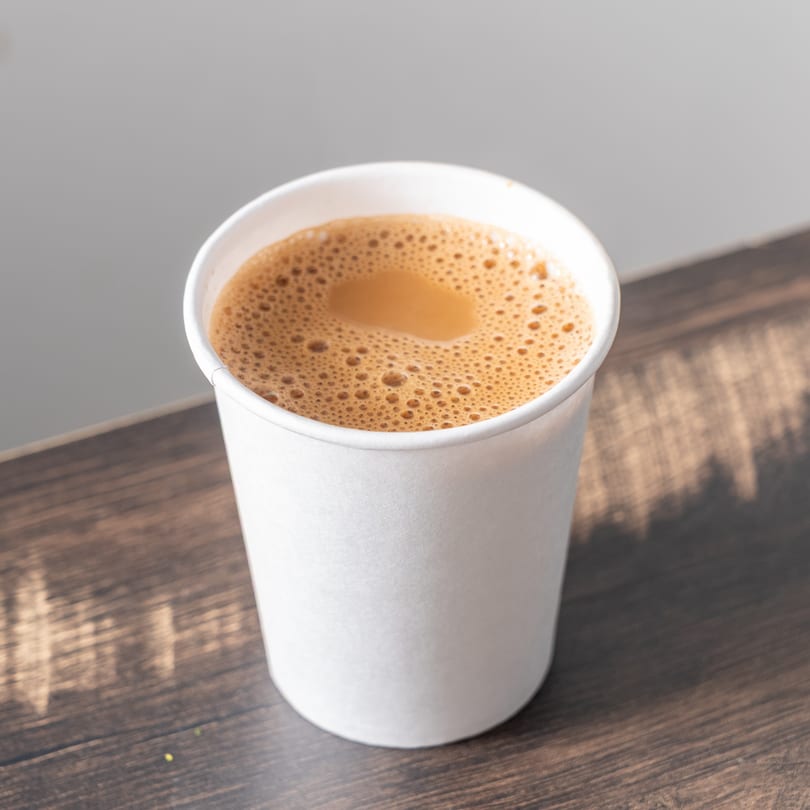 A white paper cup filled with frothy, light brown chai tea sits on a dark wooden surface.