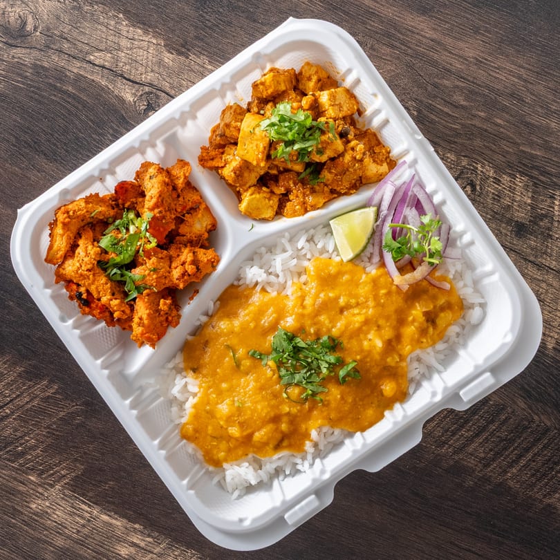 Indian takeout meal with chicken tikka, paneer, rice, dal, lime, and red onion in a white container.
