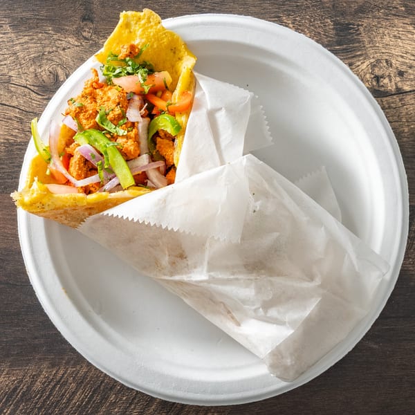 A chicken tikka roll, wrapped in paper, on a white plate atop a wooden table.