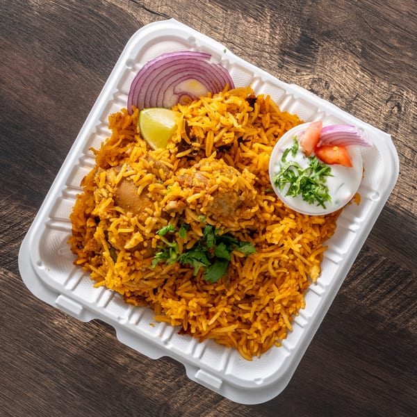 Chicken biryani with cilantro, red onions, lime, and raita in a white takeout container.