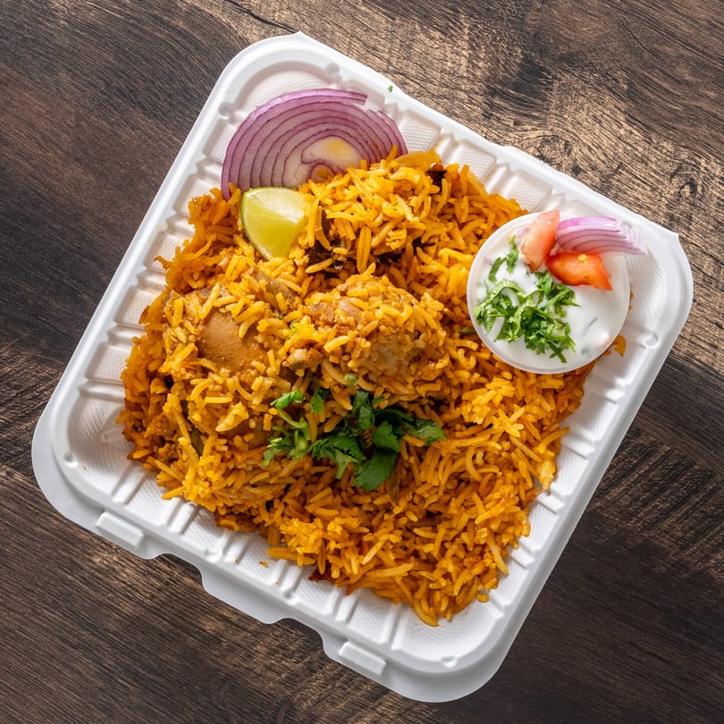 Chicken biryani with cilantro, red onions, lime, and raita in a white takeout container.