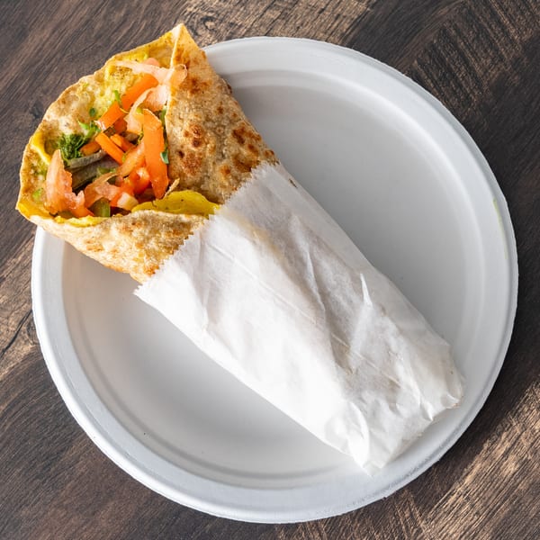 A colorful wrap filled with vegetables and meat, partially wrapped in white paper, on a white plate.
